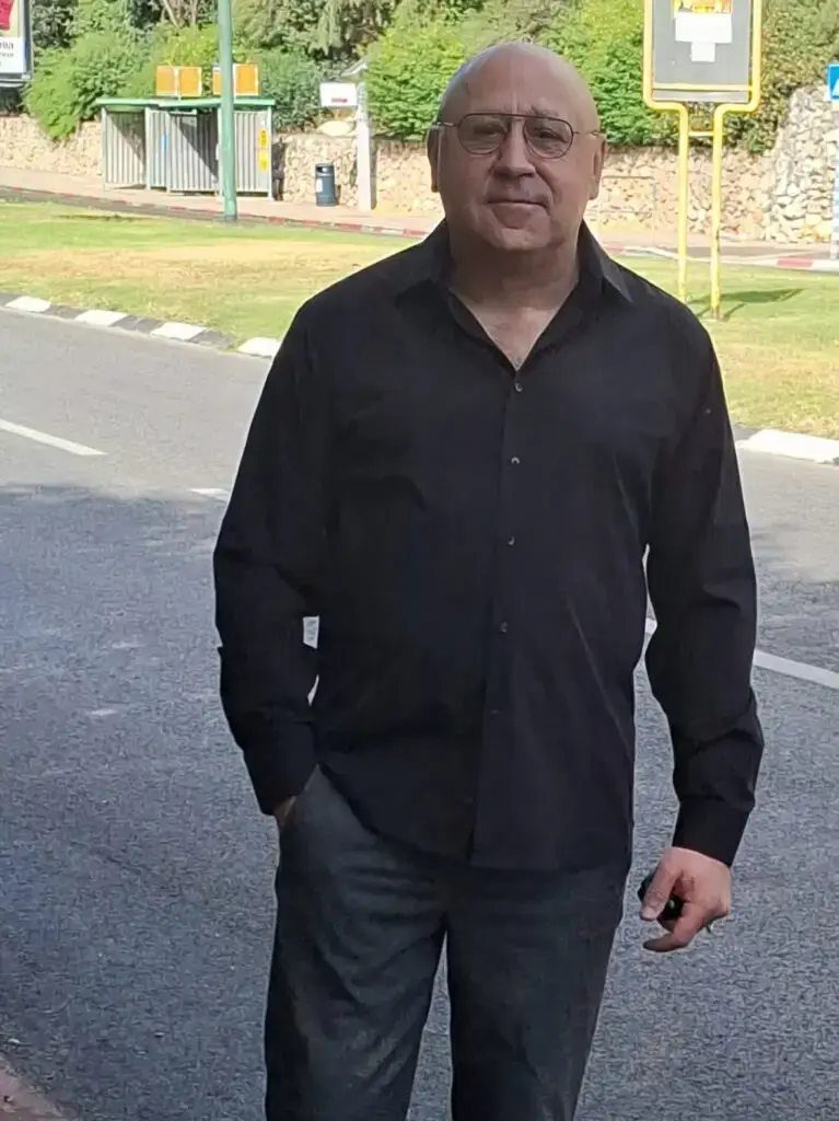 A man in a black shirt and glasses standing outdoors with one hand in his pocket.