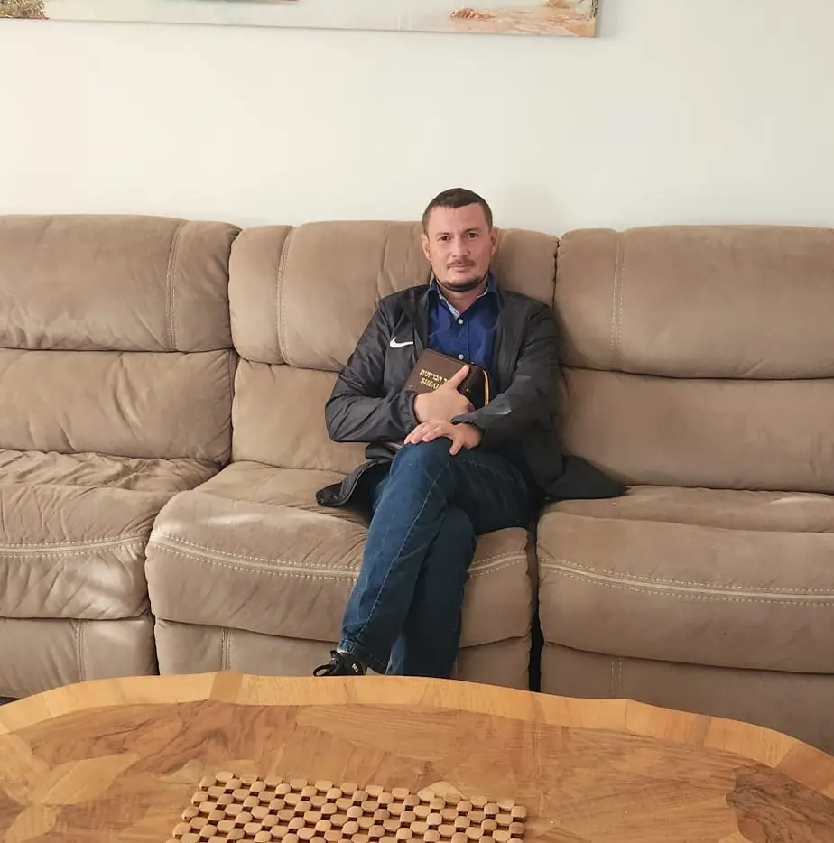 A man holding a book while sitting on a couch with a wooden coffee table in front of him.