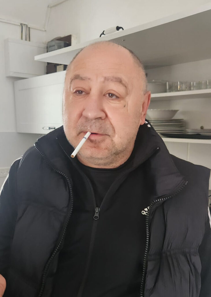 A man with a cigarette in his mouth, wearing a black vest, standing in a kitchen.