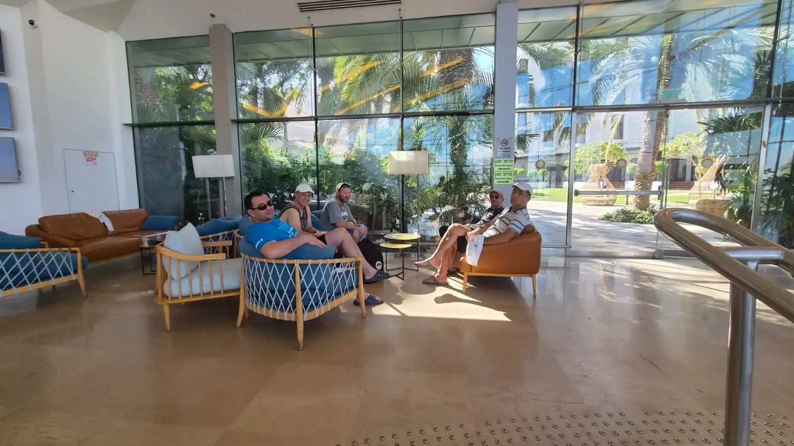 A group of men sitting together on stylish lounge chairs in a bright sunlit resort lobby for Dead Sea rest and renewal.